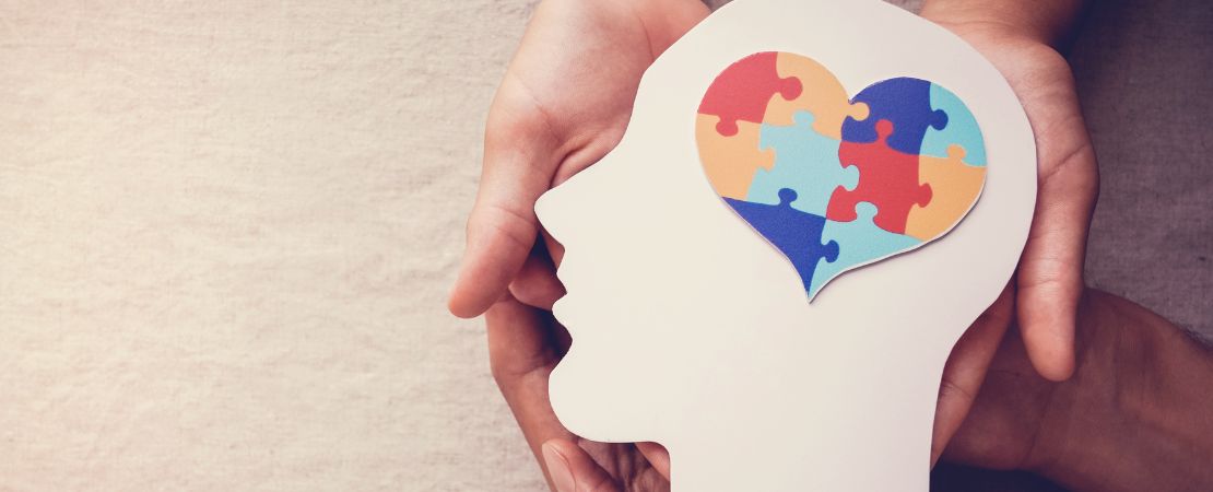 The image shows a pair of hands gently holding a cut-out silhouette of a human head in profile. Inside the head, there is a heart shape filled with colorful jigsaw puzzle pieces in red, blue, yellow, and green, symbolizing cognitive or emotional complexity, possibly referencing mental health or autism awareness.